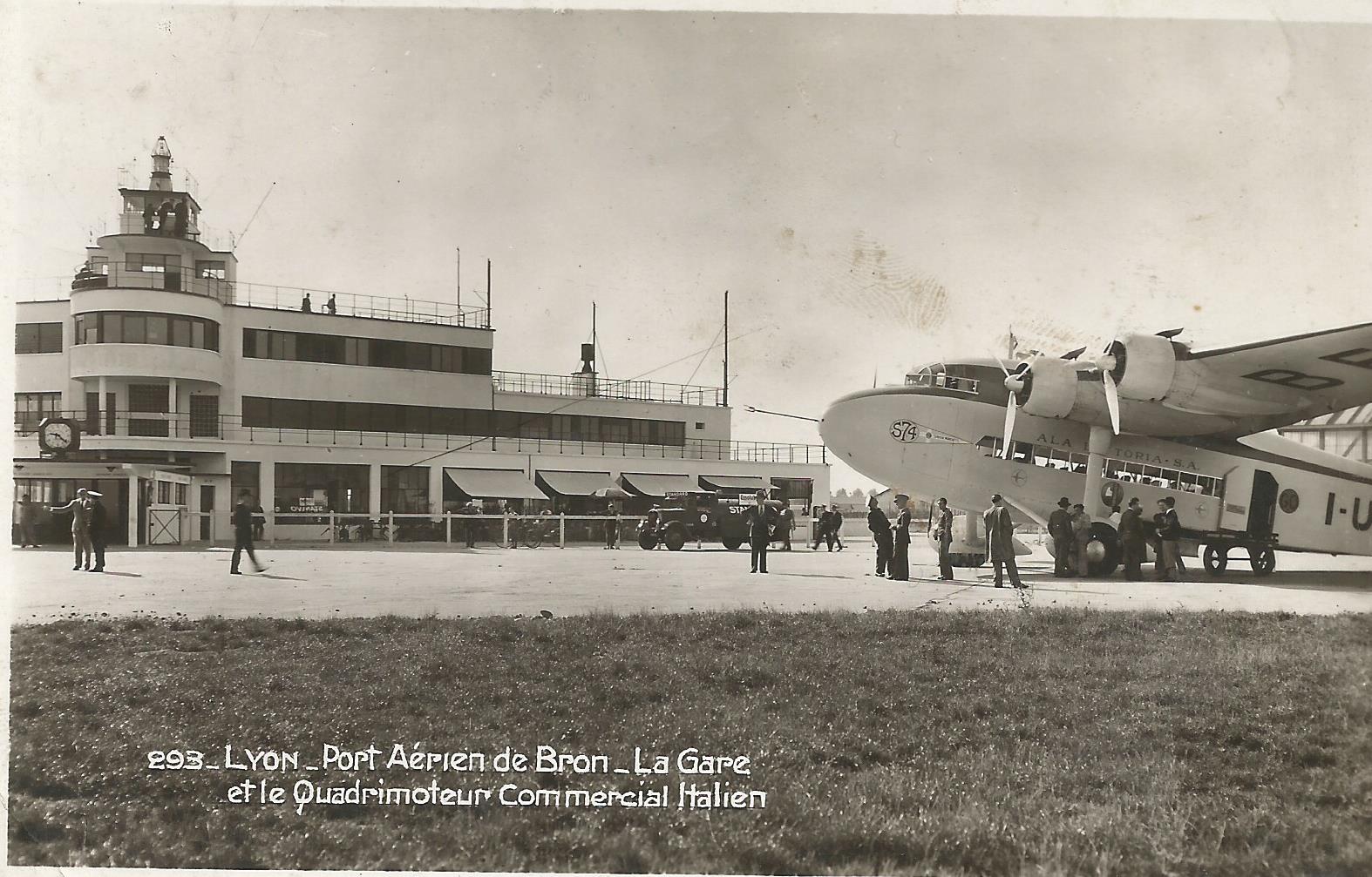 Carte postale représentant une photographie de l'aéroport de Bron en 1938, présentant, vu depui sles pistes, le bâtiment principal avec la tour de contrôle, et un avion devant avec du personnel au sol.