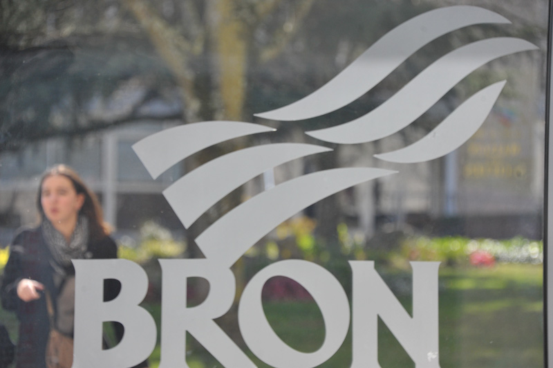 Logo de la Ville de Bron sur une vitrine, avec le square Grimma et fond. Une jeune femme passe dans en arrière plan.