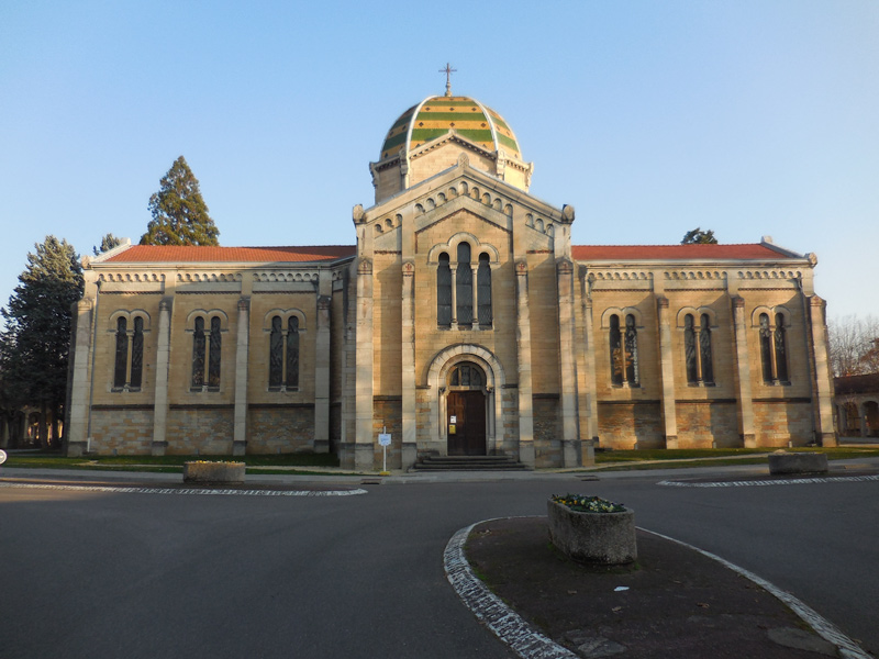 Une grande chapelle présentant une architecture en pierre, avec des murs comportant plusieurs fenêtres romanes, une porte en bois sculpté au centre, et un toit en forme de dôme, orné de tuiles dessinant des lignes horizontales vertes et jaunes, avec une croix discrète sur le sommet.