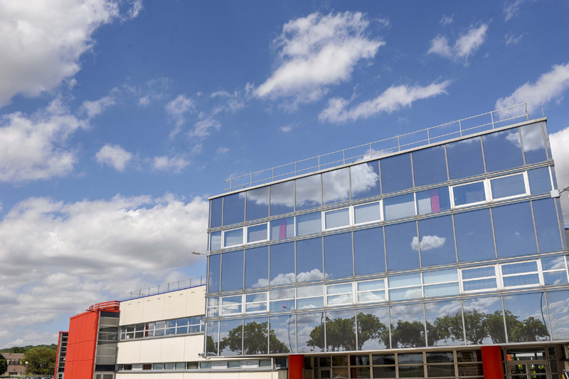 Dans le Parc du Chêne, un parc d'activités, vue sur un bâtiment tout en architecture de miroirs et fenêtres, avec un pan de mur blanc au second plan et une extension en rouge au troisième plan. D'autres bâtiments hébergeant des activités professionnelles se devinent au fond à gauche avec quelque sarbres, tandis que tout le haut de l'image est occupé par un ciel bleu parsemé de nuages. Des arbres et le ciel se reflètent dans les miroirs du bâtiment au premier plan.