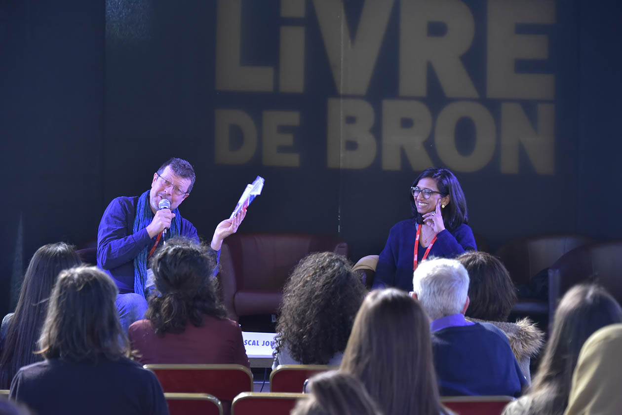 Deux auteurs, un homme et une femme, échangent assis sur une scène devant une salle remplie de spectateurs assis et nou storunant le dos. L'atmosphère est sombre et les deux personnes sont éclairées avec des tons bleutés et mauves. Les écritures "Fête du Livre de Bron " en lettres capitales et en blanc sont projetés sur le fond de la scène. La scène se déroule pendant la Fête du Livre 2017.