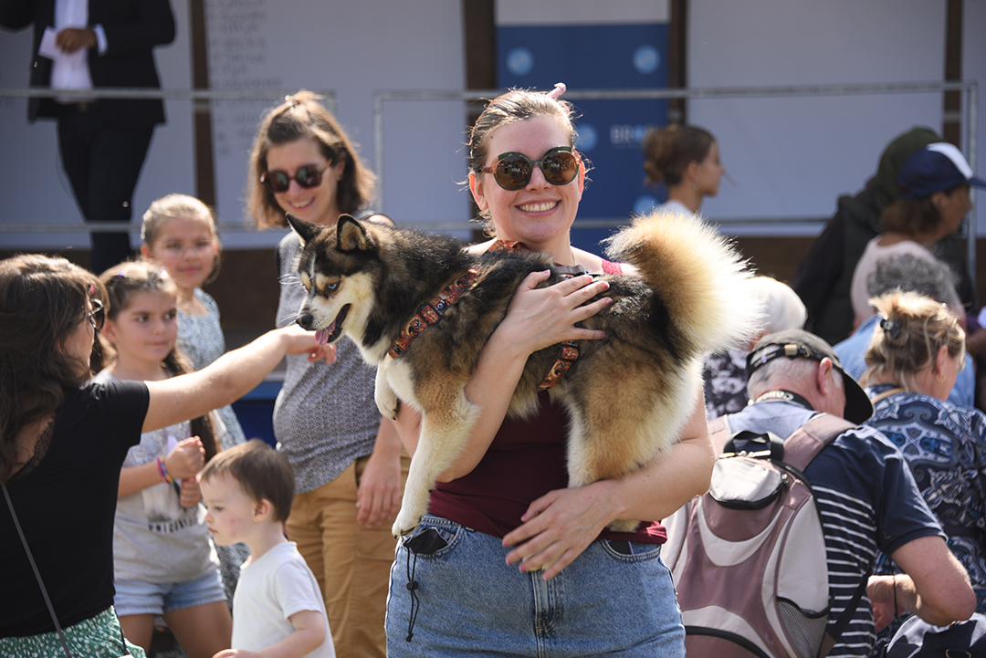 Une habitante sourit en portant son chien dans les bras, un Pomsky, des habitants discutent derrière, pendant la Fête des animaux 2024.