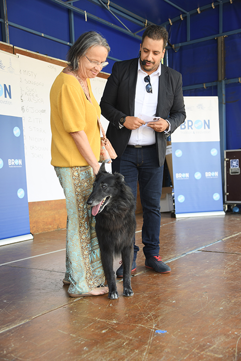 Pendant la Fête de animaux 2024, un chien noir à quattre pattes sur la scène du concours du plus beau chien, tenu en laisse par sa maîtresse que l'on voit derrière lui. L'animateur du concours est à côté.