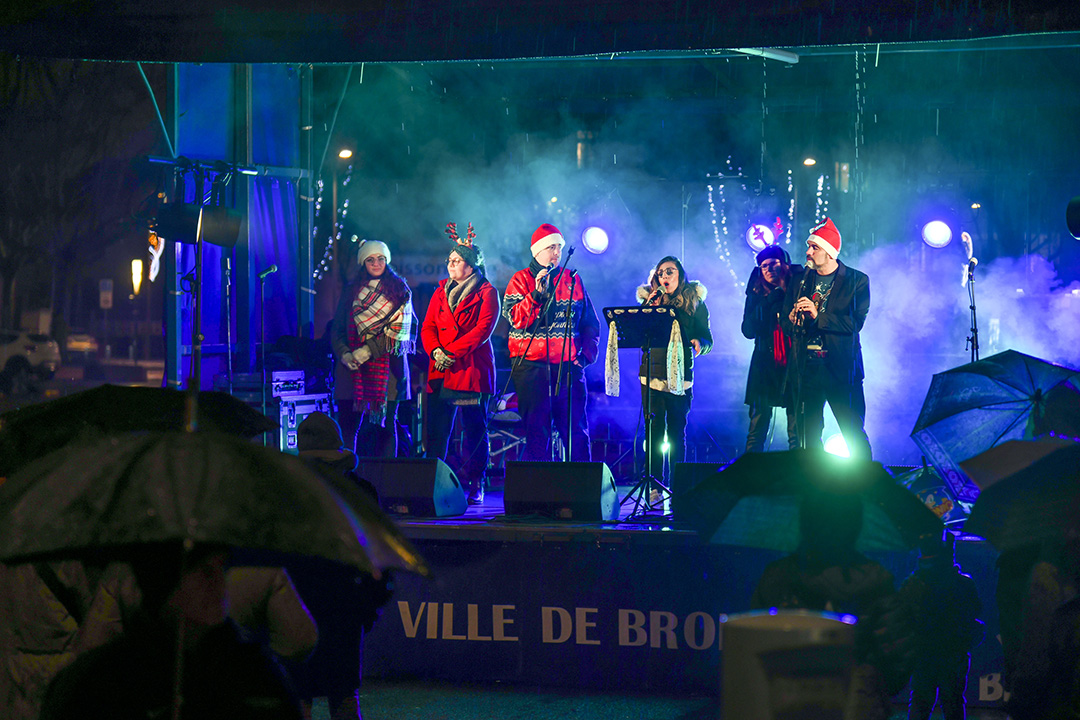 Des chanteurs sur la scène installée place de la Liberté à l'occasion du 8 décembre 2024. Des lumières et des fumigènes les entourent, certains sont grimés avec des accessoires de Noël (bonnets de père Noël et vestes rouges).