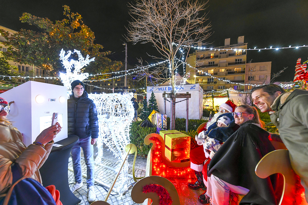 Pendant le marché de Noël 2024 de la Ville de Bron, en nocturne, des habitants se font photographier sur un traineau de Père Noël factice au milieu des cadeaux, décorations et illuminations.