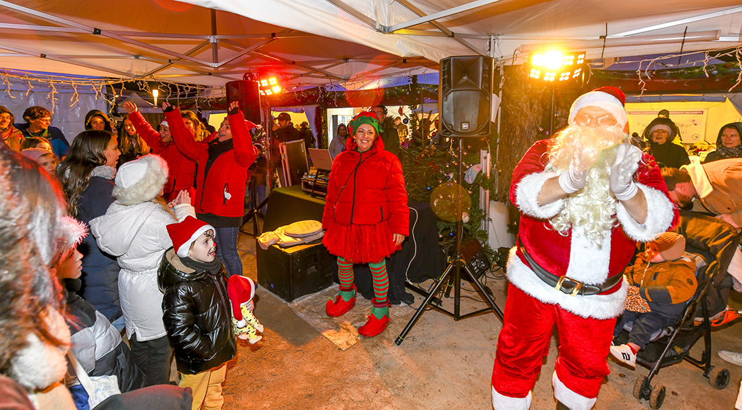 La boum du père Noël avec des enfants, le Père Noël et des personens déguisées en lutin chantent et dansent sous une large tente au marché de Noël de la Vilel de Bron en décembre 2024.