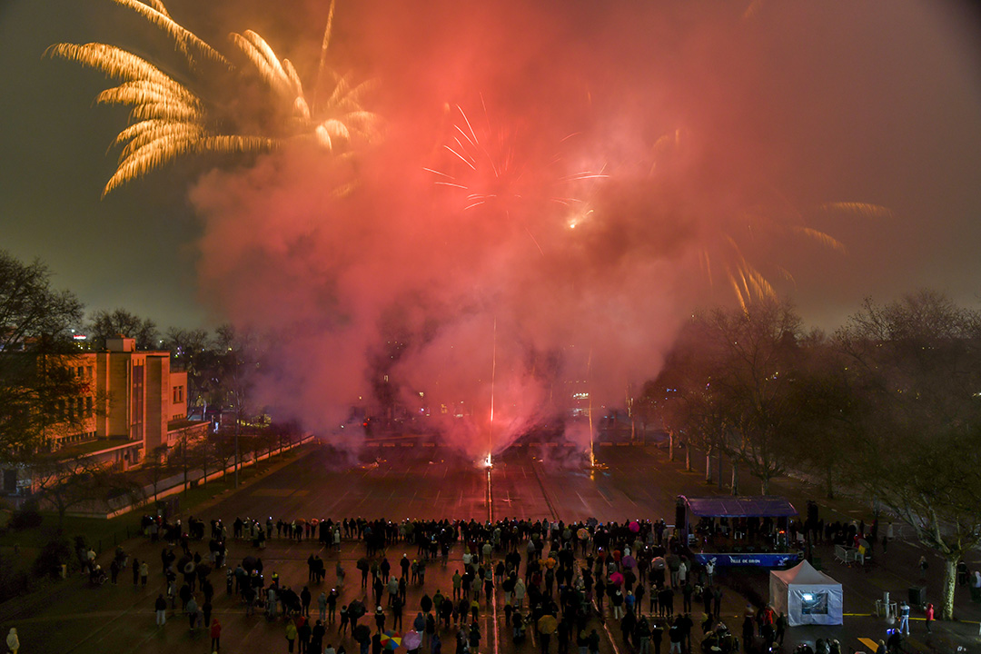 Un feu d’artifice illuminant le ciel nocturne avec des éclats dorés et rouges, une foule de personnes observe le spectacle sur le grand parking de la place de la Liberté à travers une épaisse fumée colorée.