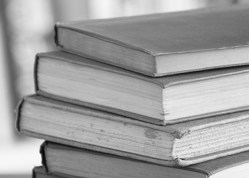 Photographie en noir et blanc représentant un empilement de livres anciens. Le fond de l'image est flou, quelques livres sont tâchés.