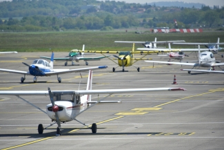 Une dizaine de petits avions de tourisme sont garés sur le tarmac de l'aéroport sous un grand soleil. On devine une plaine herbue et une colline basse arborée au fond de la photo.