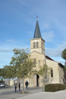 Eglise Saint Denis de Bron, architecture en pierre avec un clocher au dessus de la porte. Deux petits arbres devant le bâtiment et trois personnes passent devant l'église, porte ouverte. On devine le cimetière communal sur la gauche de l'église et une haie avance vers la place à droite de l'église.