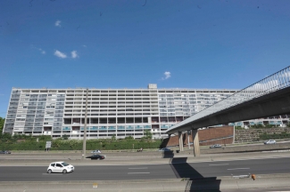Barre d'immeuble de l'UC 1, vue depuis le côté ouest du boulevard Périphérique, que l'on voit au premier plan, avec des vehicules circulant dessus, la passerelle qui l'enjambe et se dirige vers l'UC1. Le Bâtiment UC1 n'existe plus aujourd'hui et de nouveaux projets urbanistiques vont le remplacer.