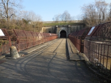 Entrée du Fort de Bron, un pont métallique, avec des garde-corps en fer rouge, menant directement à une grande entrée de tunnel sombre encadrée de pierres.