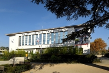Hôtel de Ville de Bron, un bâtimentrectangulaire peint en blanc avec une façade présentant une grand ebaie de plusieurs fenêtres en son centre, la devise Liberté - Egalité - Fraternité sur le fronton, un drapeau français flotte au dessu de la porte, au centre en bas de la façade. Un cèdre lance ses branche au premier plan et une place arborée au second plan est reliée au bâtiment par quelques marches.