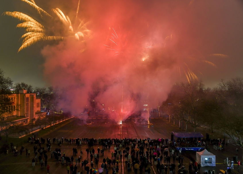Un feu d’artifice illuminant le ciel nocturne avec des éclats dorés et rouges, une foule de personnes observe le spectacle sur le grand parking de la place de la Liberté à travers une épaisse fumée colorée.
