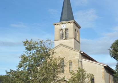 Eglise Saint Denis de Bron, architecture en pierre avec un clocher au dessus de la porte. Deux petits arbres devant le bâtiment et trois personnes passent devant l'église, porte ouverte. On devine le cimetière communal sur la gauche de l'église et une haie avance vers la place à droite de l'église.