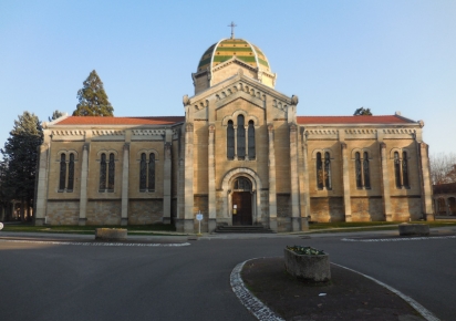 Une grande chapelle présentant une architecture en pierre, avec des murs comportant plusieurs fenêtres romanes, une porte en bois sculpté au centre, et un toit en forme de dôme, orné de tuiles dessinant des lignes horizontales vertes et jaunes, avec une croix discrète sur le sommet.