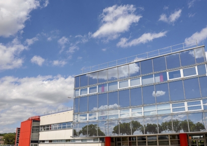 Dans le Parc du Chêne, un parc d'activités, vue sur un bâtiment tout en architecture de miroirs et fenêtres, avec un pan de mur blanc au second plan et une extension en rouge au troisième plan. D'autres bâtiments hébergeant des activités professionnelles se devinent au fond à gauche avec quelque sarbres, tandis que tout le haut de l'image est occupé par un ciel bleu parsemé de nuages. Des arbres et le ciel se reflètent dans les miroirs du bâtiment au premier plan.