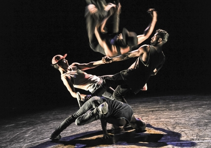 Quatre danseurs de Hip Hop effectuent une chorégraphie acrobatique façon pyramide humaine sur l ascène de l'espace Albert Camus à l'occasion du festival Karavel en 2016. Le fond et le sol sont noirs et aucun décor ne les entourent.