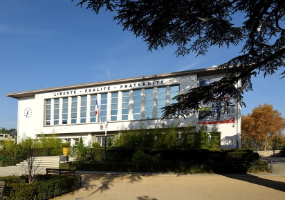 Hôtel de Ville de Bron, un bâtimentrectangulaire peint en blanc avec une façade présentant une grand ebaie de plusieurs fenêtres en son centre, la devise Liberté - Egalité - Fraternité sur le fronton, un drapeau français flotte au dessu de la porte, au centre en bas de la façade. Un cèdre lance ses branche au premier plan et une place arborée au second plan est reliée au bâtiment par quelques marches.