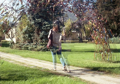 Une femme semblant semi-transparente, comme un fantôme, probablement à cause d'une double exposition photographique, portant un casque audio et marchant sur un sentier entouré d'herbe et d'arbres.