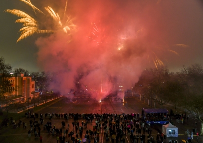 Un feu d’artifice illuminant le ciel nocturne avec des éclats dorés et rouges, une foule de personnes observe le spectacle sur le grand parking de la place de la Liberté à travers une épaisse fumée colorée.