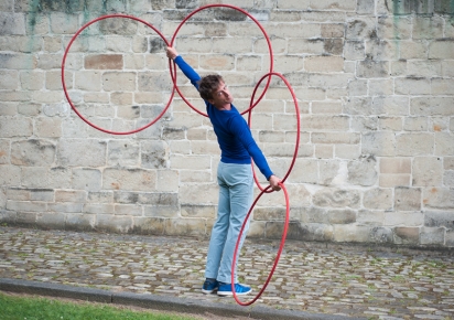 Photographie en couleur d'un artiste, de dos, à moitié tourné vers nous, qui présente quatre cerceaux rouges emmêlés entre ses deux bras étendus au maximum, devant un mur de pierre, sur un sol pavé..