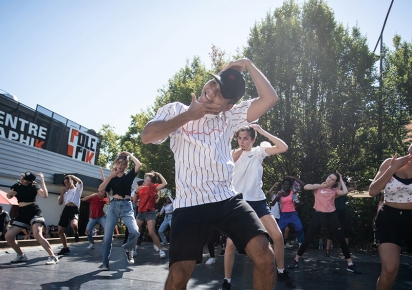 Photographie en couleurs d'une troupe de danseurs de Hip Hop en pleine chorégraphie en plein air, sur le parvis du Pôle Pik, que l'on voit dans le fond à gauche de l'image.