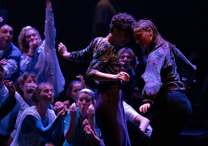 deux danseurs se tournent autour au premier plan, tandis qu'un groupe de danseurs font mine de les encourager, les éclairages sont dan sles teintes violettes, le fond de l ascène est noir